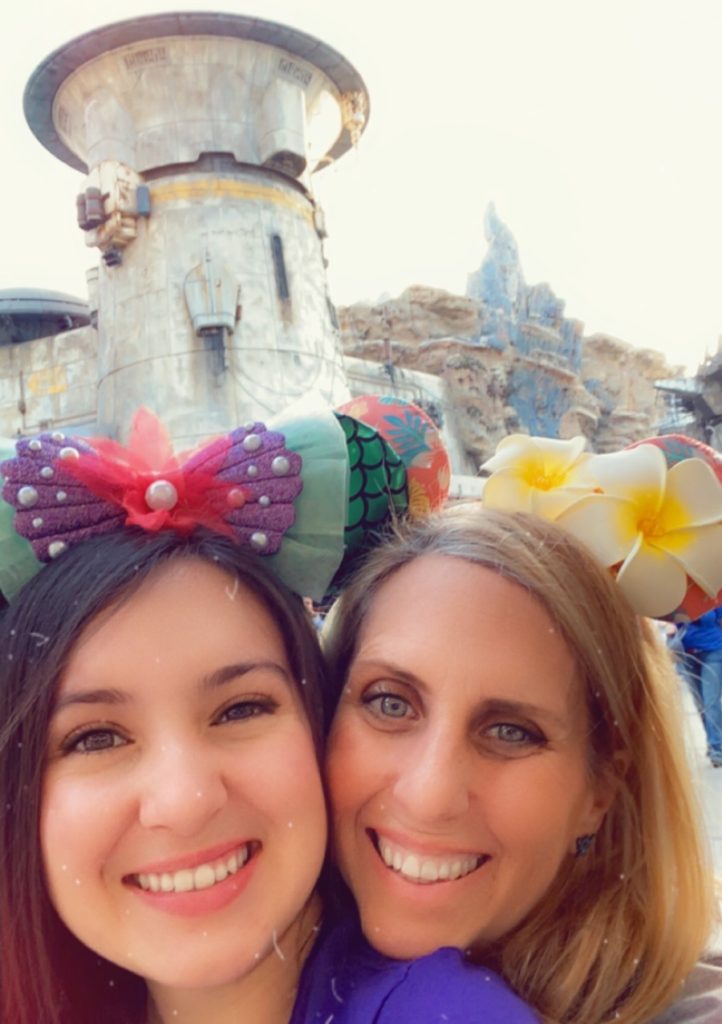 A woman on the left (Jay) is wearing The Little Mermaid themed mouse ears and smiling, and a blonde woman on the right (Shell) is smiling and wearing Hawaii themed mouse ears.