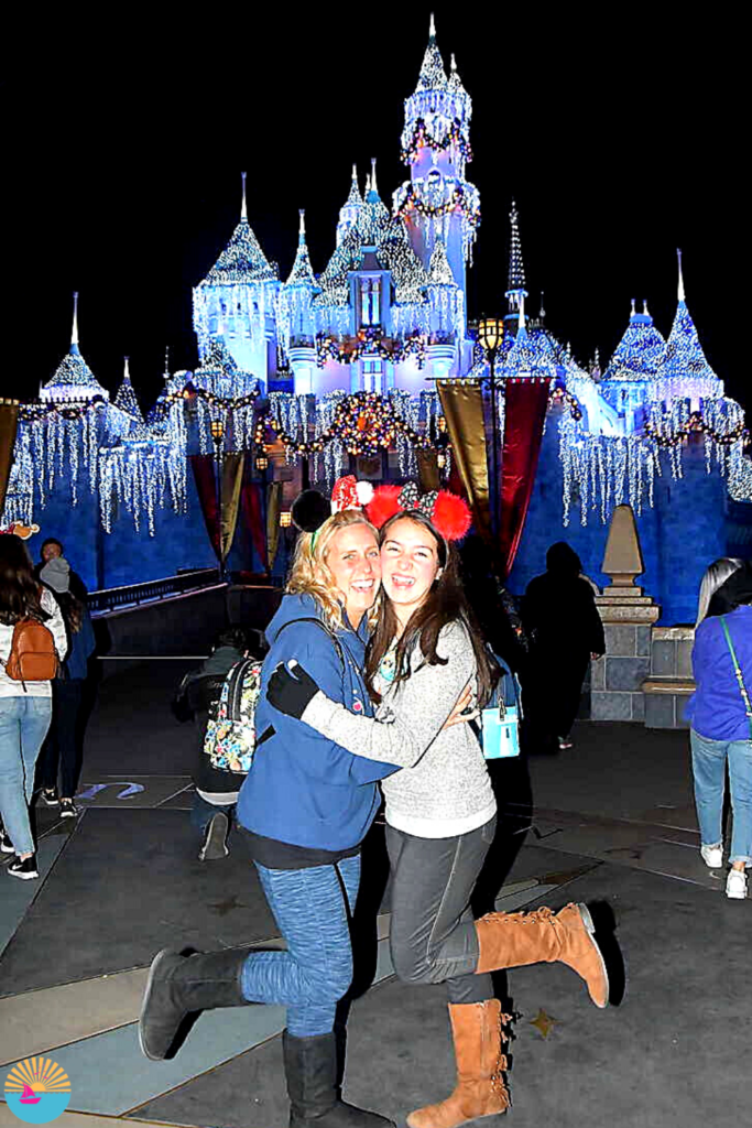 Jay and Shell hug and laugh in front of the holiday decorated Sleeping Beauty's Castle at Disneyland. Both women have one leg lifted and Holiday mouse ears on their heads.