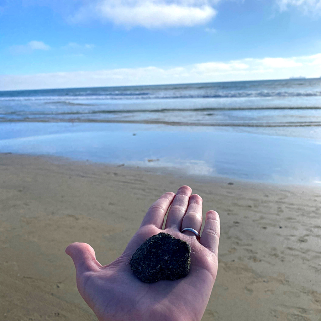 In the palm of a woman's hand sits a black rock, with a background of the sand and calm ocean waves. 