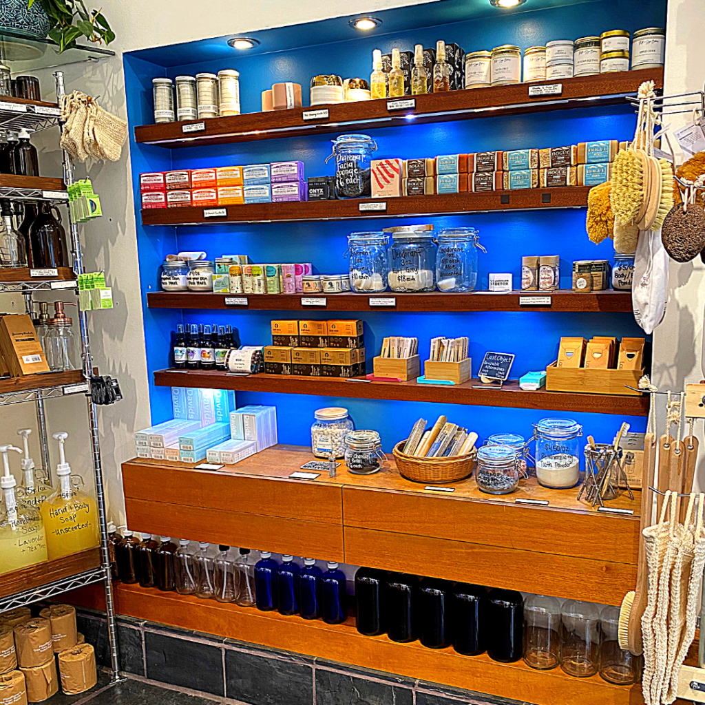A wall of blue paint has wooden selves full of soap bars, brushes, perfume in glass jars, and bamboo toothbrushes. There is an assortment of glass spray bottles available for sale underneath. 