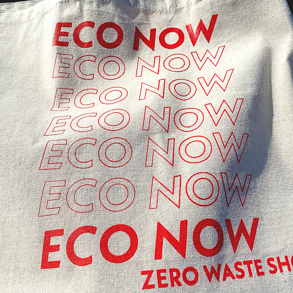 A reusable bag repeats "Eco Now" across the front of it in red writing. It is a zero waste alternative to plastic bags.