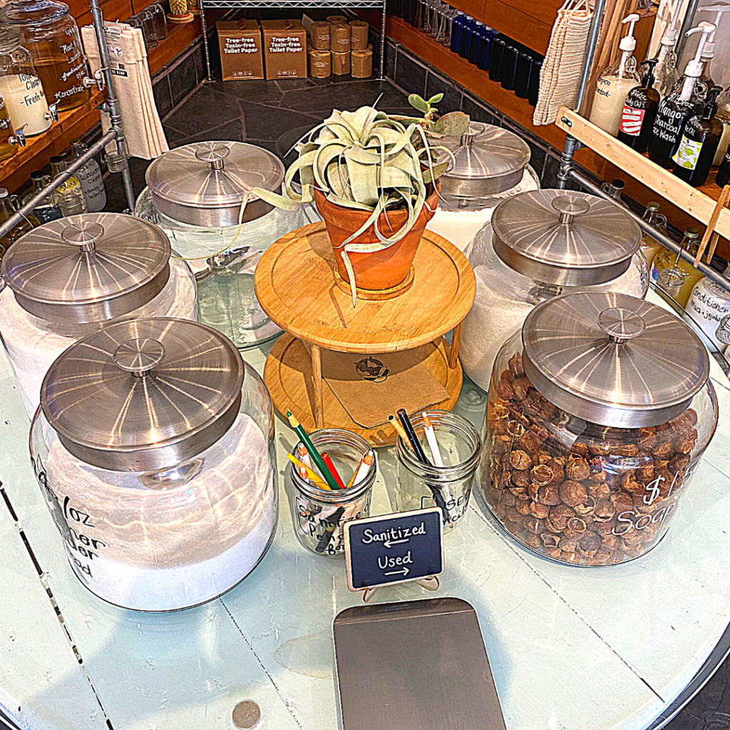 A table of painted white wood with a layer of glass over it displays jars of eco-friendly products such as laundry detergent. There is a succulent plant in the middle. Additionally, there is a scale in the front. On the left side is a glass of pencils marked "sanitized" and on the right is a glass jar of pencils marked "used".